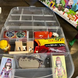 Image of a plastic multi-bin container that has different snacks and treats in it to create a homemade advent calendar. The plastic bin is open showing the following items inside: beef jerky, individual candy bar, roll of stickers, paw patrol mini figure, Pusheen magnets.