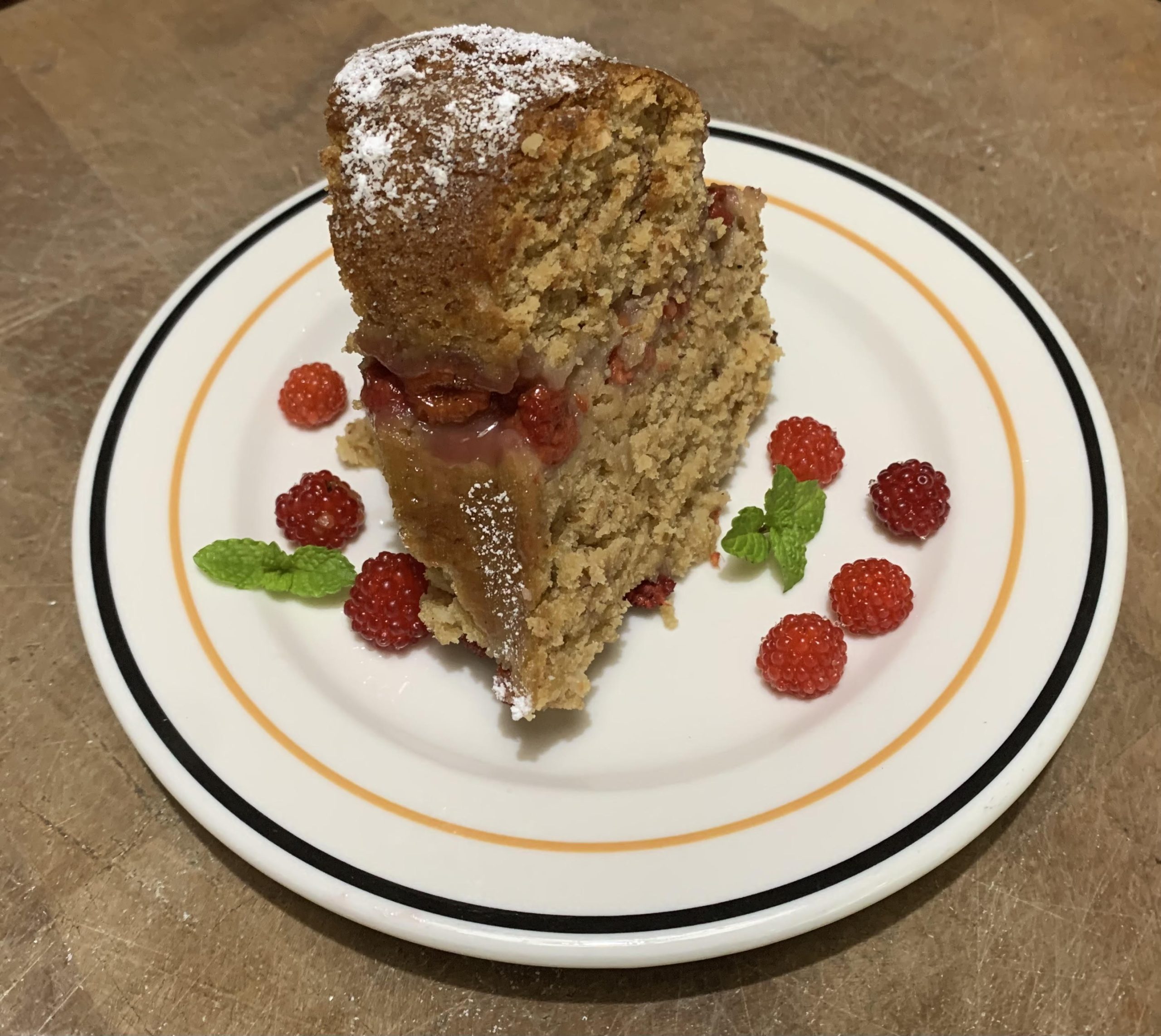 Slice of vegan raspberry cake on a plate with fresh raspberries scattered on the plate around it. Powdered sugar is sprinkled on the top.
